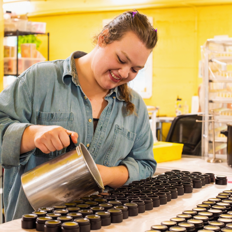 A woman in a denim shirt smiles while pouring liquid from a metal pitcher into many small black jars arranged on a table in a bright workshop with yellow walls.