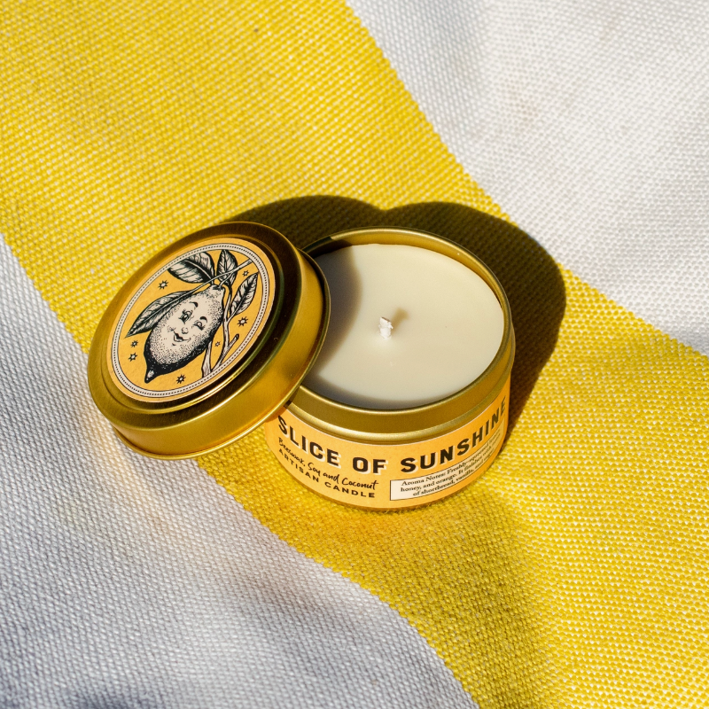 An open gold metal candle tin labeled 'SLICE OF SUNSHINE' with cream-colored wax inside, placed on a yellow-and-white striped fabric background.