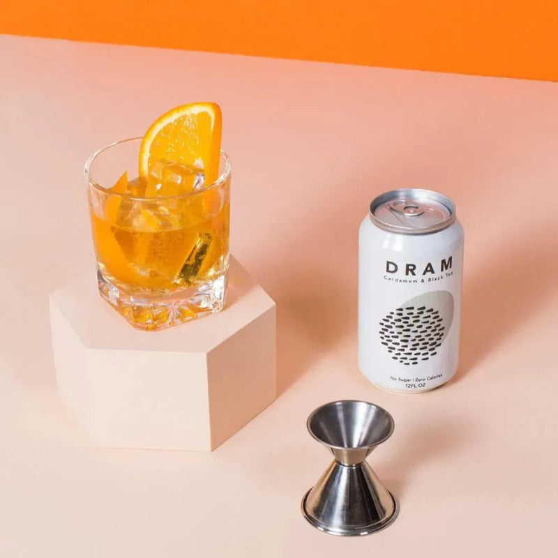Glass of orange cocktail with a slice of orange, DRAM can, and jigger on a peach and orange background