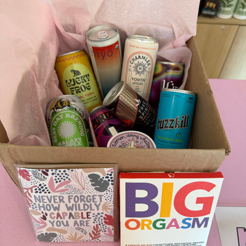 Box with beer cans and a colorful book titled 'BIG ORGASM' on a pink surface.