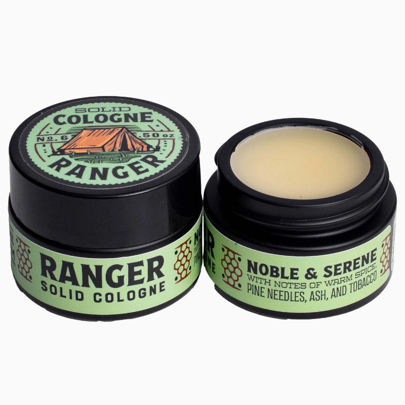 Two black cosmetic jars of solid cologne on a white background, one closed and one open showing the balm inside. The labels are light green with black text and feature a small orange tent illustration.