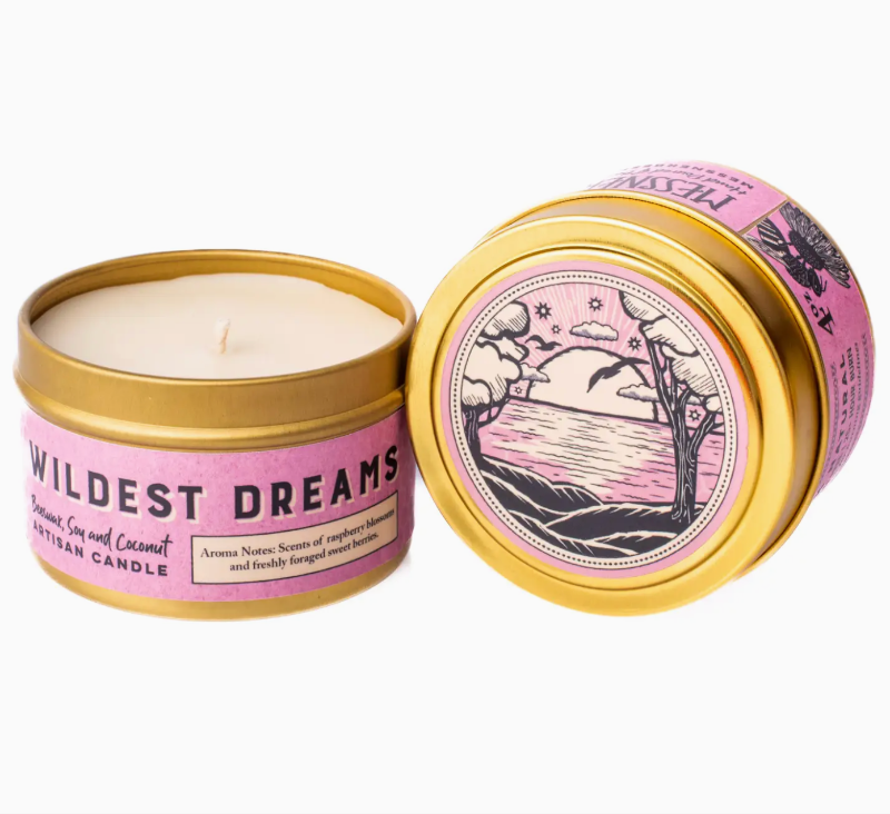 Two tins of a candle on a plain white background, one open showing the cream-colored wax and wick, and one closed with a pink and gold label that reads 'WILDEST DREAMS' and shows a moonlit landscape illustration.
