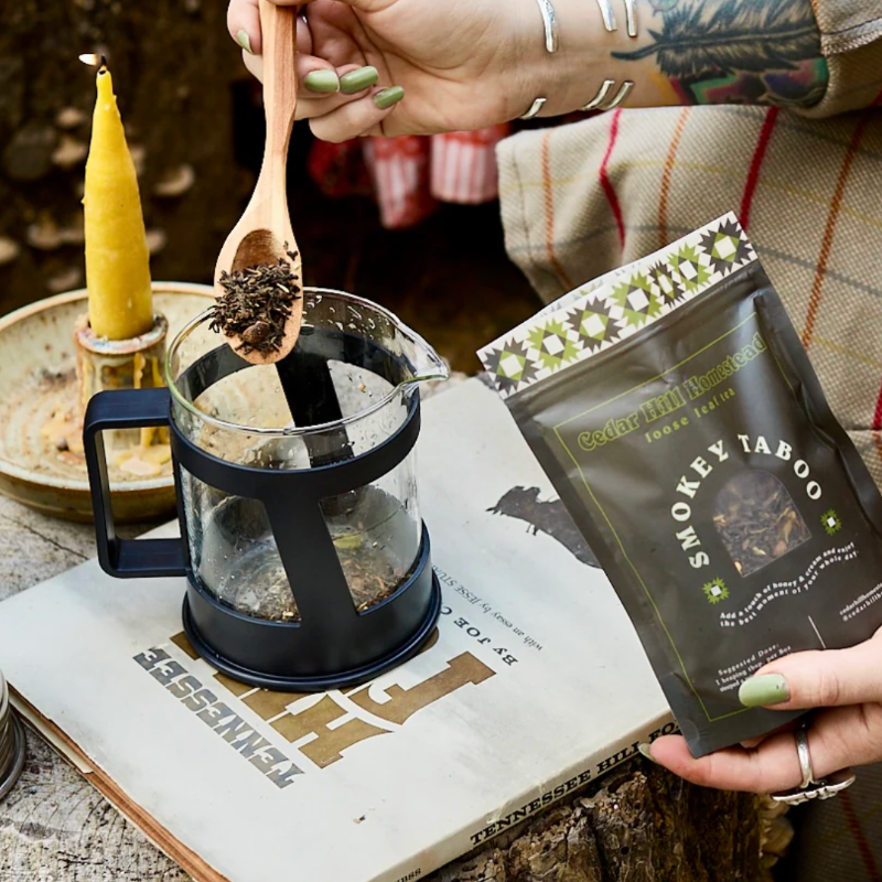 Person preparing a cup of smoky flavored tea with a wooden spoon and a package of Smokey Taboo tea.
