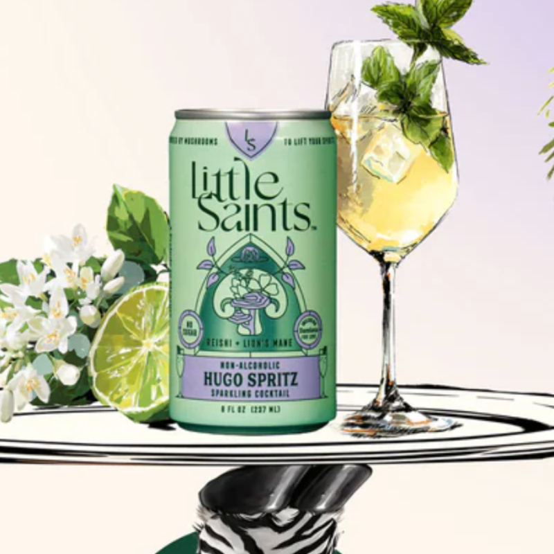 A can of Little Saints Hugo Spritz sparkling cocktail is displayed on a reflective surface next to a filled wine glass with mint and a lime wedge, against a soft pastel background.