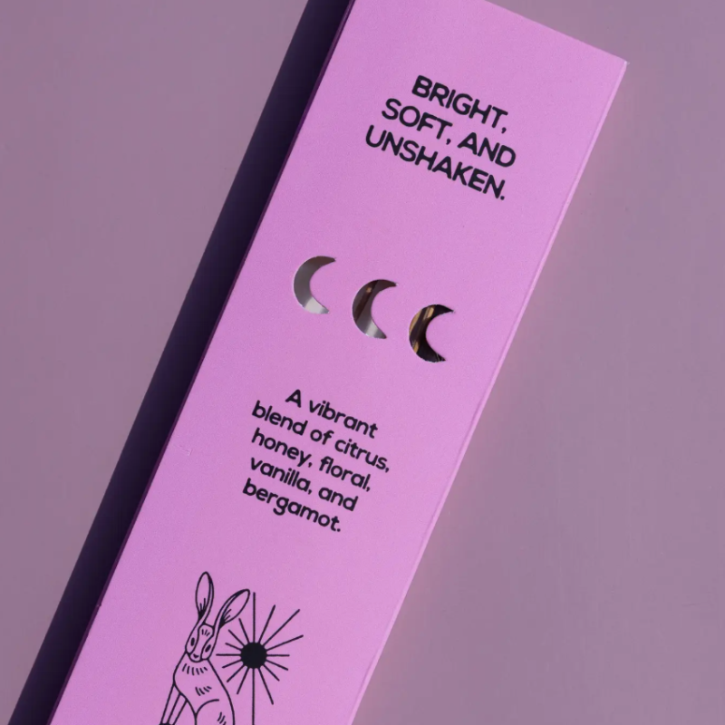 A close-up of a lavender-pink product box with black text reading 'BRIGHT, SOFT, AND UNSHAKEN.' and a scent description, featuring three moon-shaped cutouts and a rabbit illustration near the bottom.