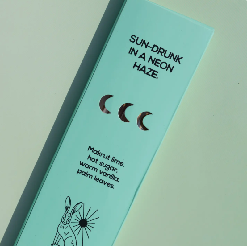 A mint green rectangular product box with black text reading 'SUN-DRUNK IN A NEON HAZE.' and flavor notes including 'Makrut lime, hot sugar, warm vanilla, palm leaves.