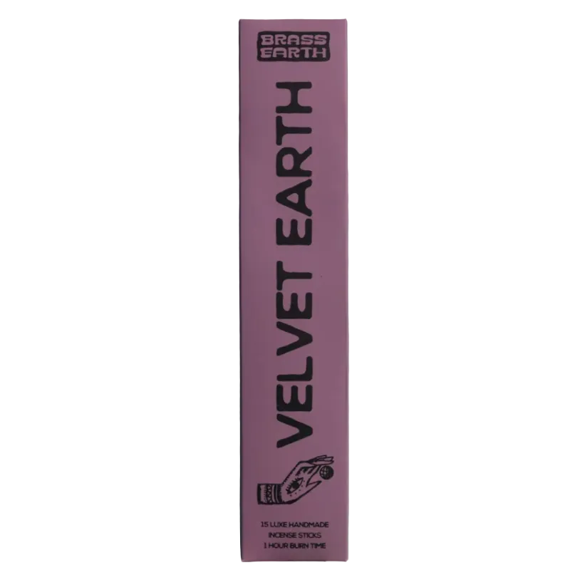 A tall, narrow pink box for Brass Earth Velvet Earth incense sticks, featuring black text and a small hand graphic near the bottom, photographed against a pale blue-gray background.