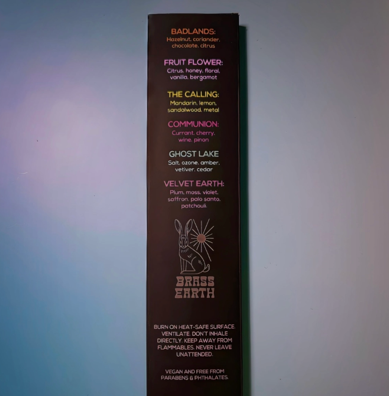 A tall, narrow dark brown cardboard display card or packaging insert for Brass Earth, featuring printed product names like Fruit Flower, The Calling, and Complimenton in small text against a blue-gray gradient background.