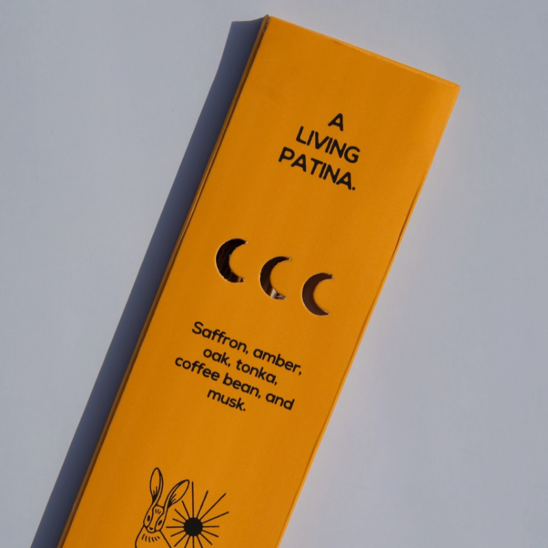 A bright yellow rectangular box for 'A LIVING PATINA.' is photographed at an angle on a light gray background.