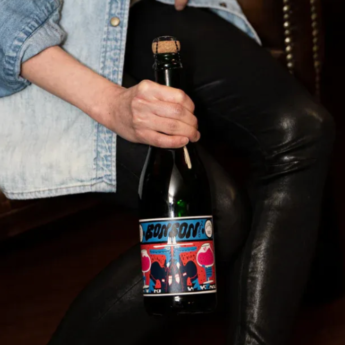 A person in a light blue denim shirt and black leather pants holds a dark glass bottle with a cork stopper and a colorful label that reads 'FATALISM'.