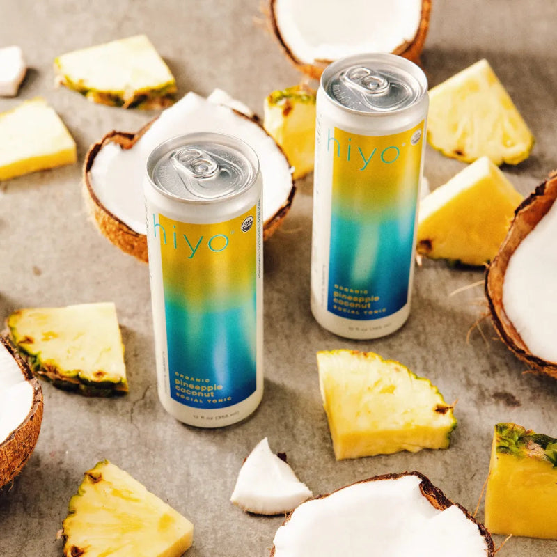 Two cans of 'hiyo' brand drink surrounded by pineapple and coconut pieces on a textured surface.